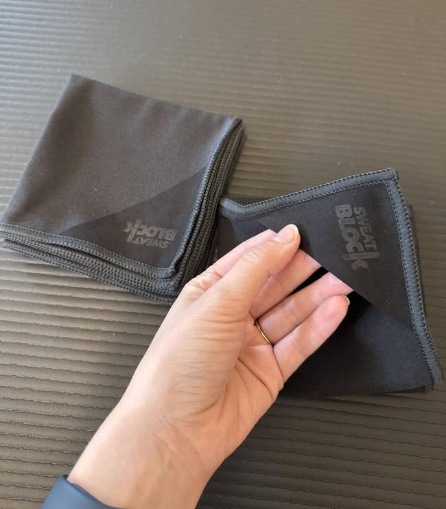 Two black microfiber cloths on a table; one is folded, and a hand is holding the corner of the other
