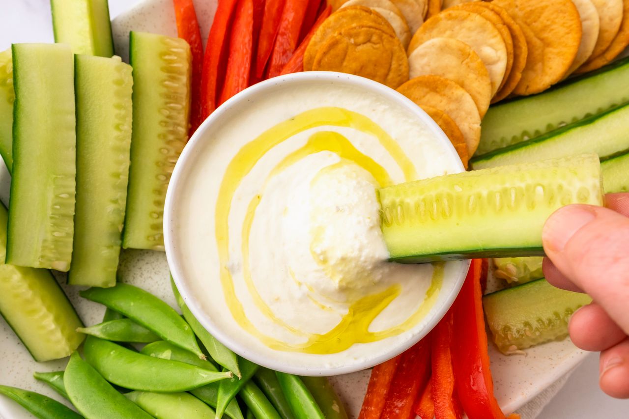A bowl of feta dip surrounded by sliced vegetables and crackers with a hand dipping a cucumber stick into it