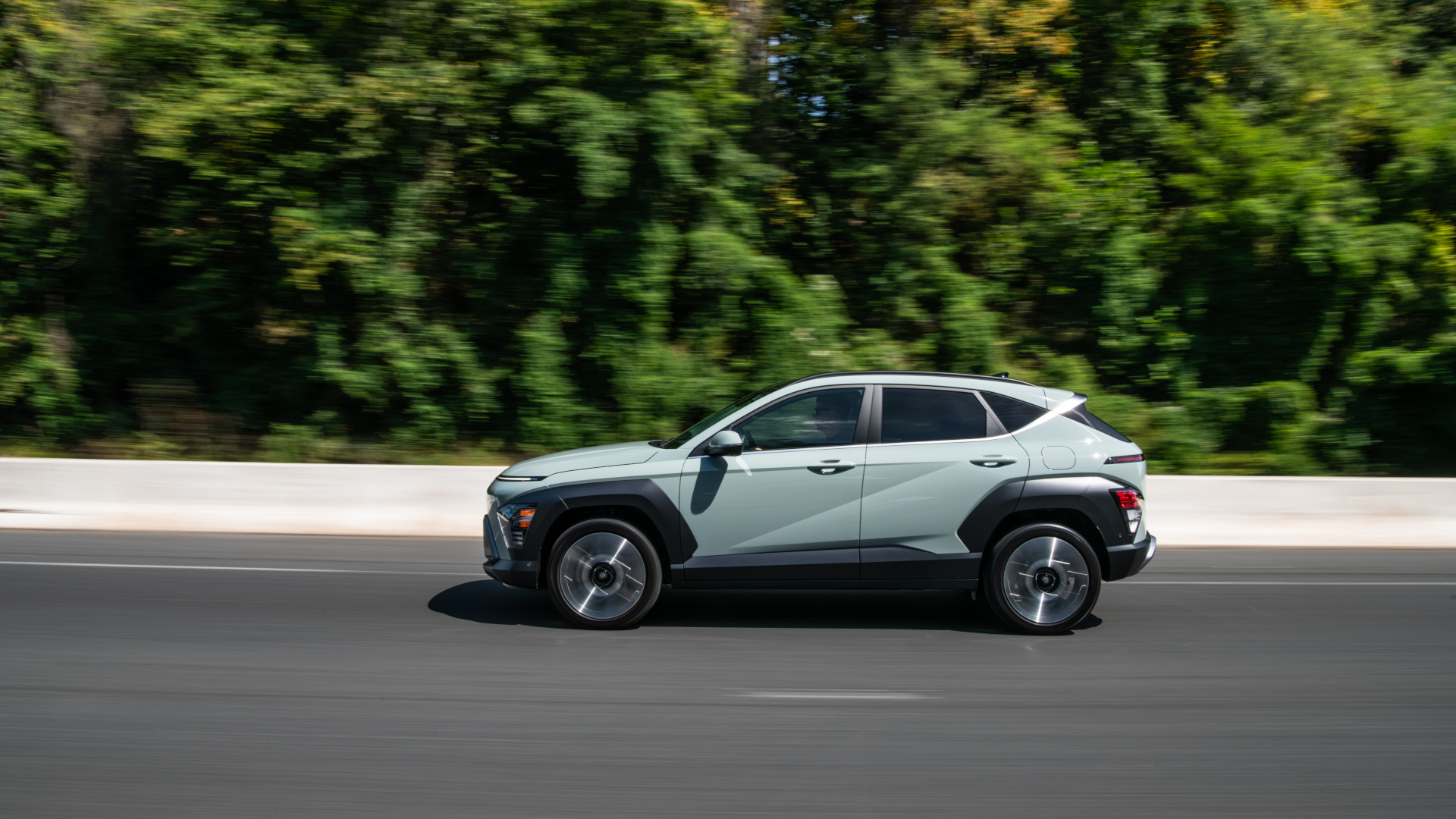 Discover A 2026 Hyundai Kona drives down a wide paved road with lush green trees alongside. it.