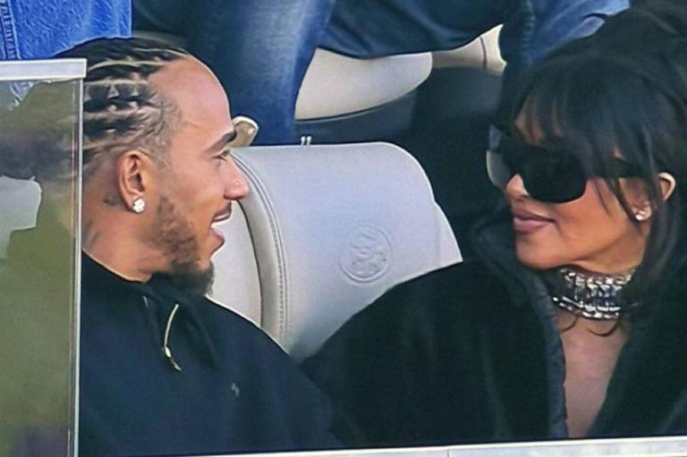 Two individuals, one male with facial hair and a distinctive hairstyle and one female wearing sunglasses and a necklace, are seated in close proximity, engaging in a conversation within a setting that appears to be a vehicle.