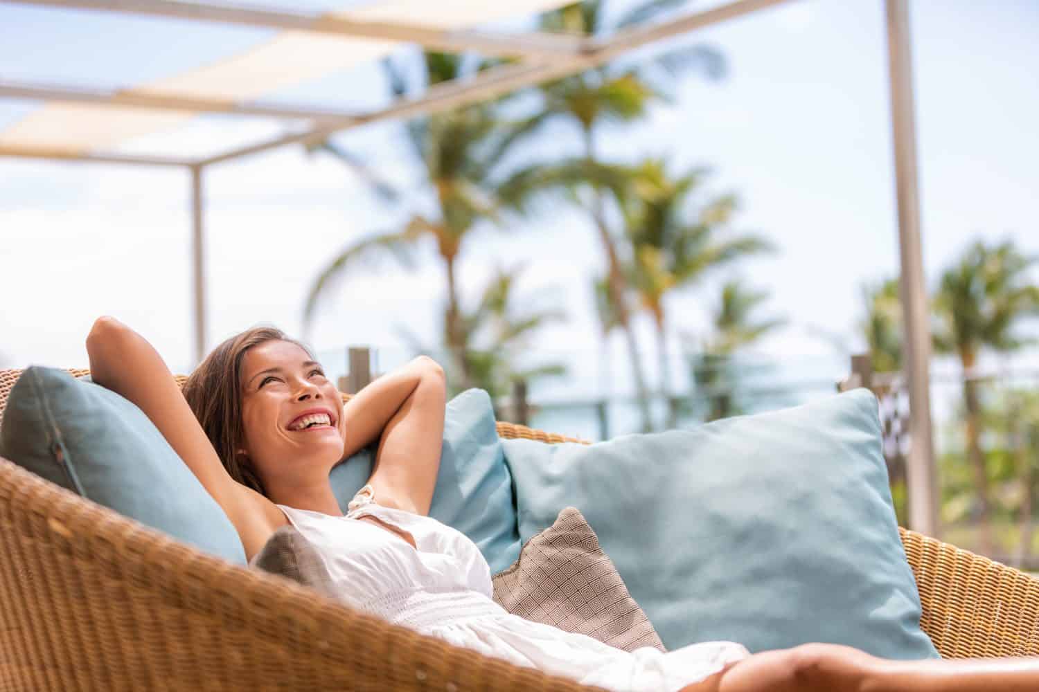 Luxury hotel home living woman relax enjoying sofa furniture of outdoor patio. Beautiful young multiracial Asian girl relaxing day dreaming for rich early retirement in getaway tropical house.