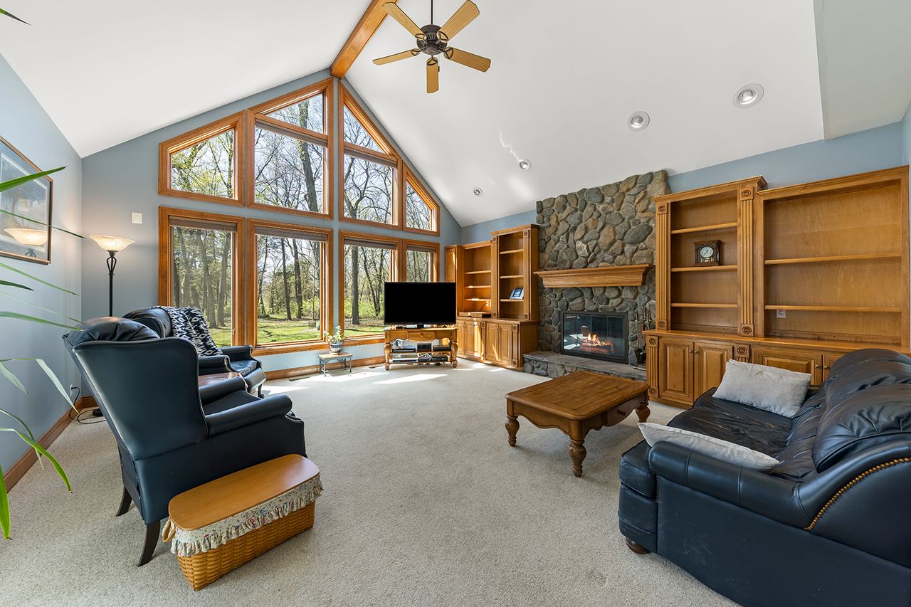 A spacious living room with a stone fireplace wooden bookshelves and large glass windows overlooking a forested area
