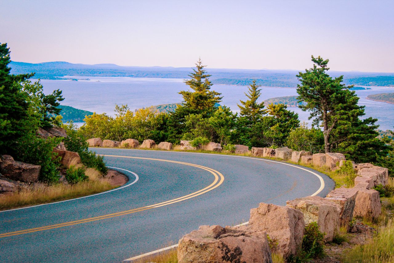 A scenic winding road bordered by rocky railings leading towards a view of dense forest and water
