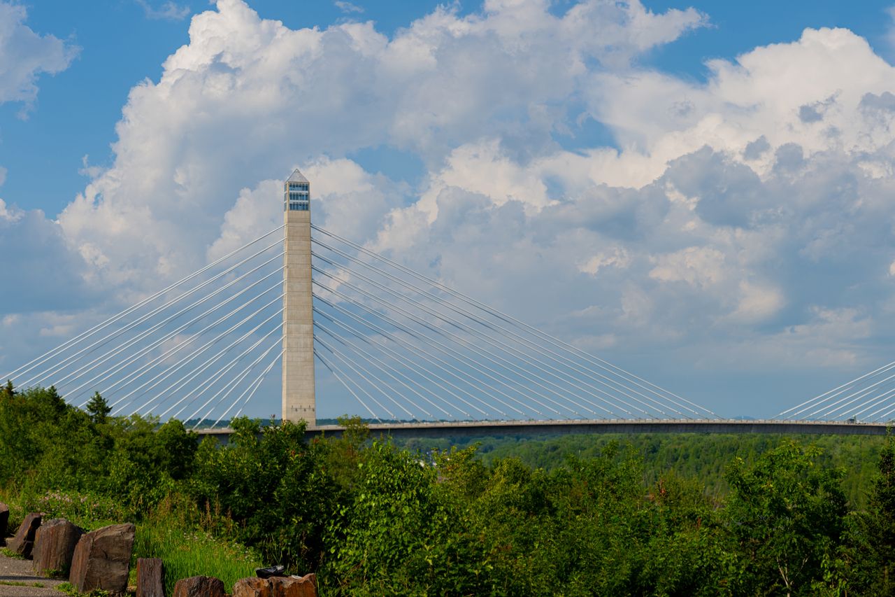 A cablestayed bridge tower extending against a backdrop of clouds and greenery