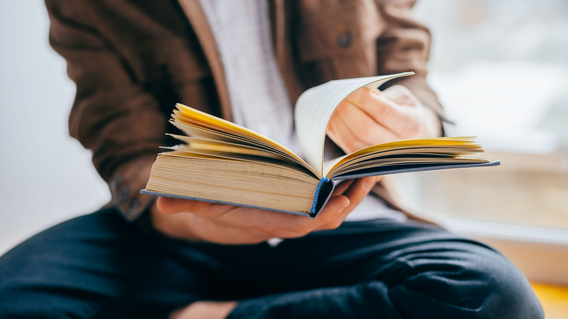 Discover a young man wearing a light brown jacket or flannel flips through the pages of an old or vintage book while seated