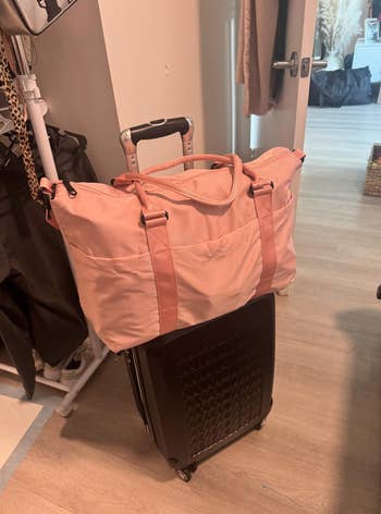 A pink travel bag with pockets is placed on top of a black rolling suitcase in a home setting, implying travel or packing