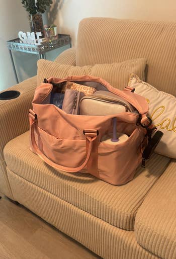 Open peach-colored diaper bag on a beige chair, filled with baby essentials like wipes, a bottle, and a changing mat
