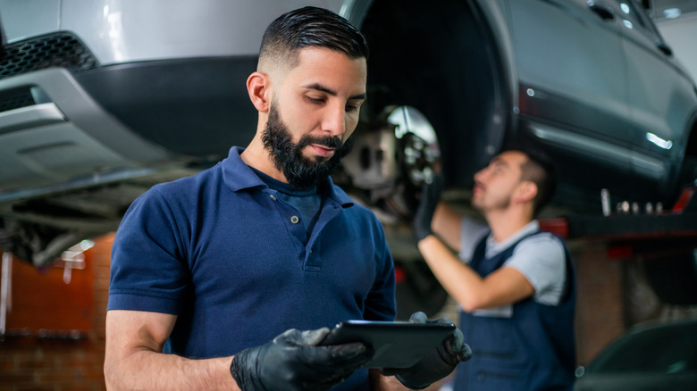 Service technician holding phone while a second technician services car's wheel well in background at auto shop