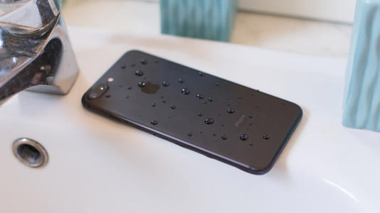 A black iPhone with specs of water on it sits on top of a white sink in what is likely a bathroom