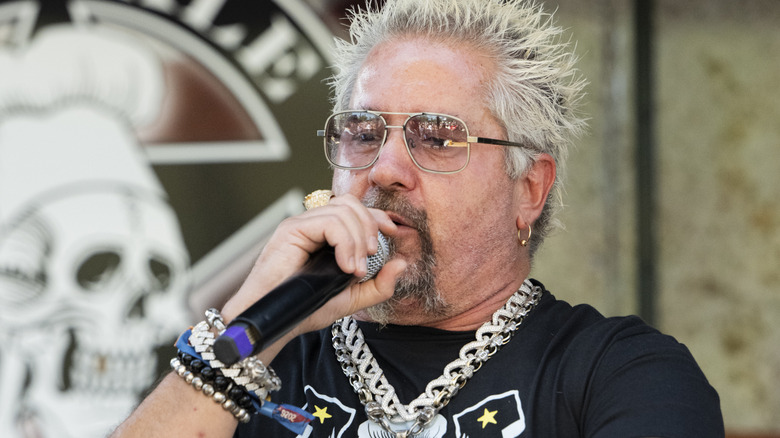 Guy Fieri is holding a microphone while speaking at an event.