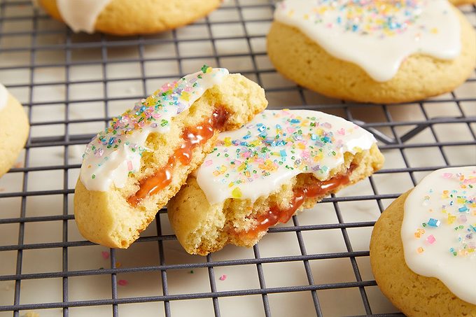 Eye level horizontal image of a jam-filled cookie broken in half, resting on a black cooling rack. The cookie halves reveal a vibrant red filling and are topped with white icing and colorful sprinkles, with additional cookies visible in the background.