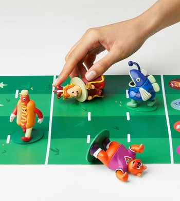 Hand moves a game piece shaped like a hot dog on a board with quirky characters