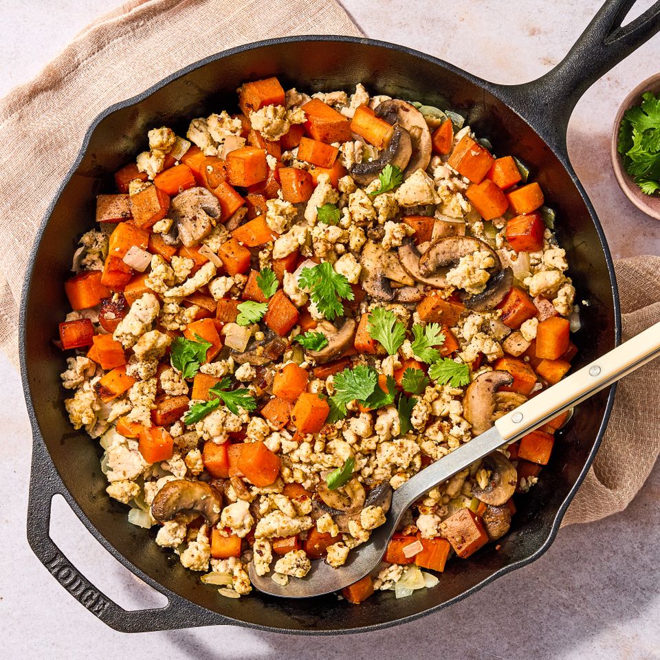 A skillet containing a mixed dish of ground turkey sweet potatoes and mushrooms garnished with cilantro with a serving spoon