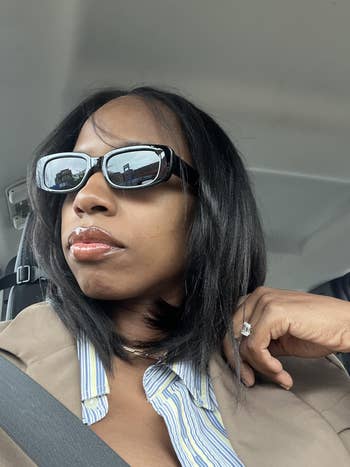 Person in car wearing sunglasses and a blazer. They have a reflective expression, with a hand resting on their chin