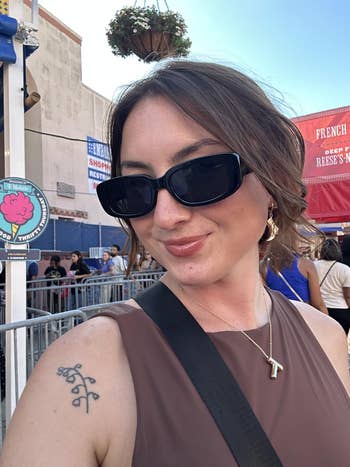 Person with short hair wearing large sunglasses and a sleeveless top, standing outdoors at a bustling market or fair