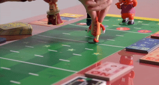 Hands move toy football players on a board game resembling a football field, suggesting a playful shopping item