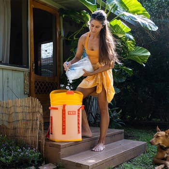 Woman pouring ice into a large YETI cooler outside a cabin. She is casually dressed and barefoot, with a dog nearby
