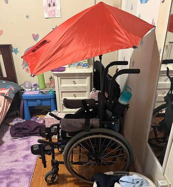 Wheelchair with a large red umbrella attached, situated in a cluttered room with clothes and furniture visible