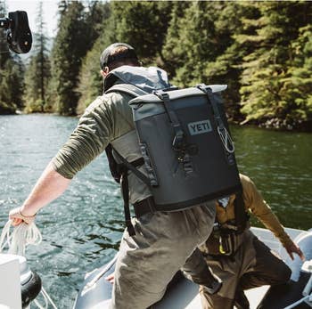 Person on a boat wearing a large Yeti backpack, preparing to step ashore in a wooded area
