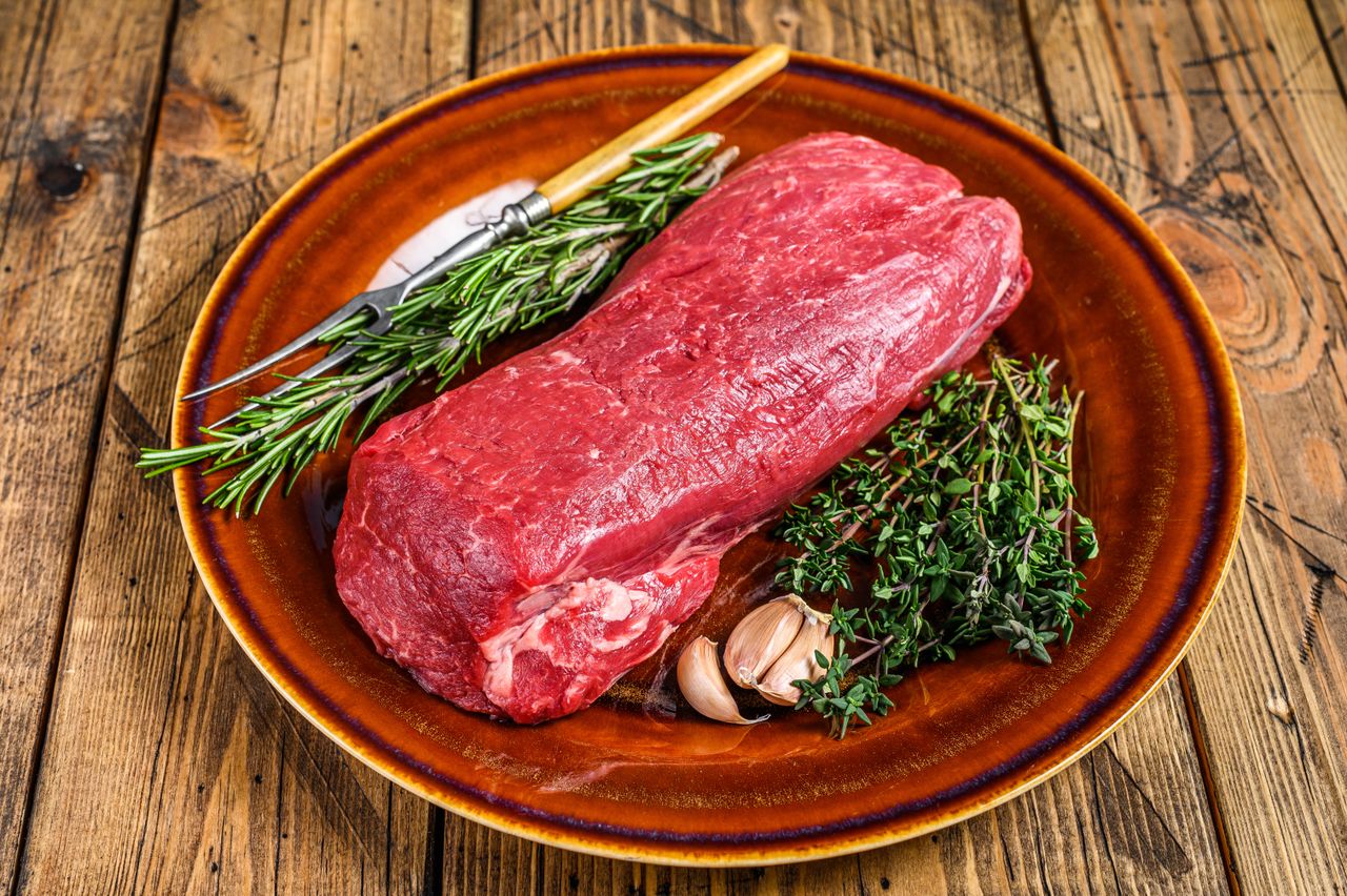 A raw filet mignon steak on a plate with herbs and garlic garnished with rosemary and thyme