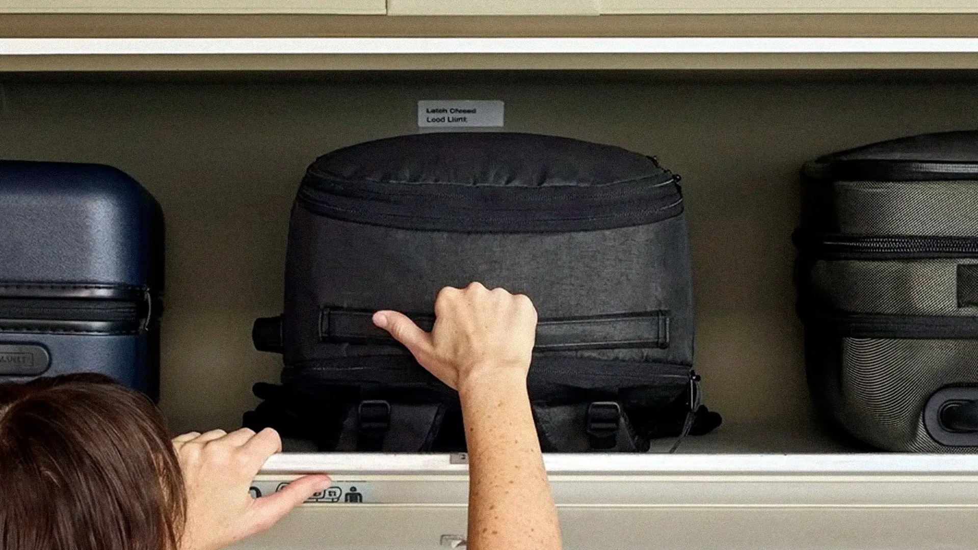 Person's hands placing a black backpack into an overhead airplane luggage compartment with other suitcases.