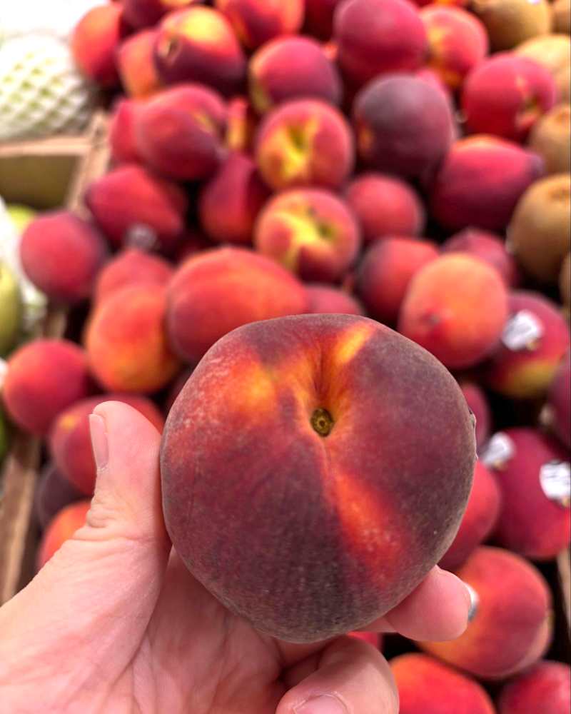 A hand holding a ripe peach in front of a display of many peaches in a market.