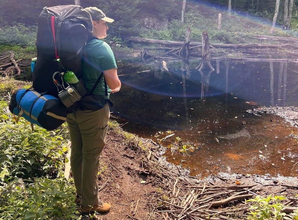 Maria Aldrich looking out onto a body of water in a forest, with a rucksack and other camping gear on her back
