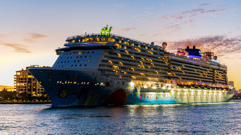 A brightly lit Norwegian Cruise Line ship illuminates a harbor at evening time with buildings in the background