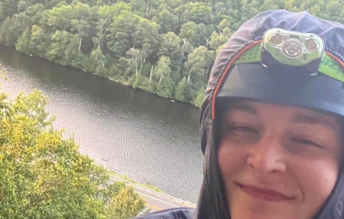 Maria Aldrich taking a selfie in outdoor gear with a view of a river and forests in the background