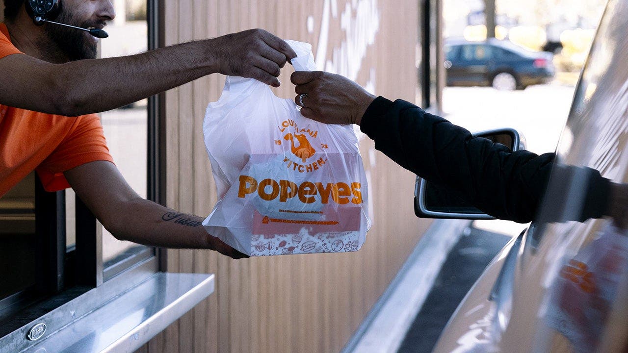 A Popeyes employee hands a customer a bag of food at a drive-thru window.
