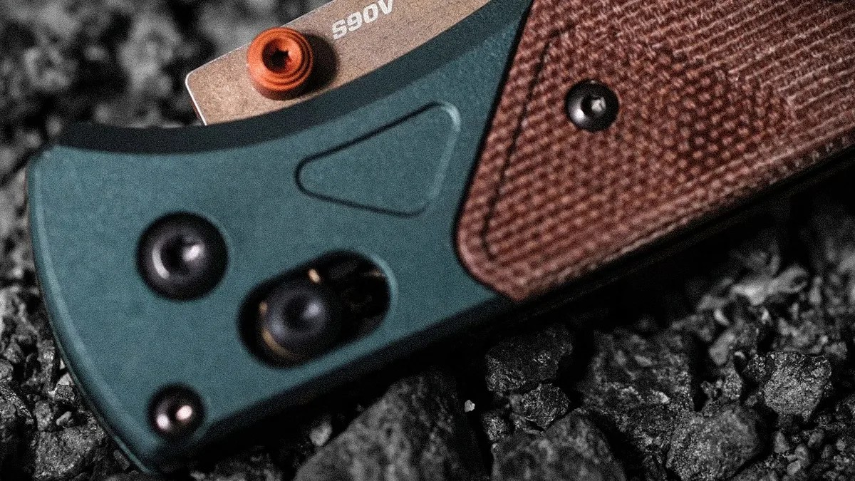 Close-up of a folding knife handle with green and brown textured grip resting on dark gravel.