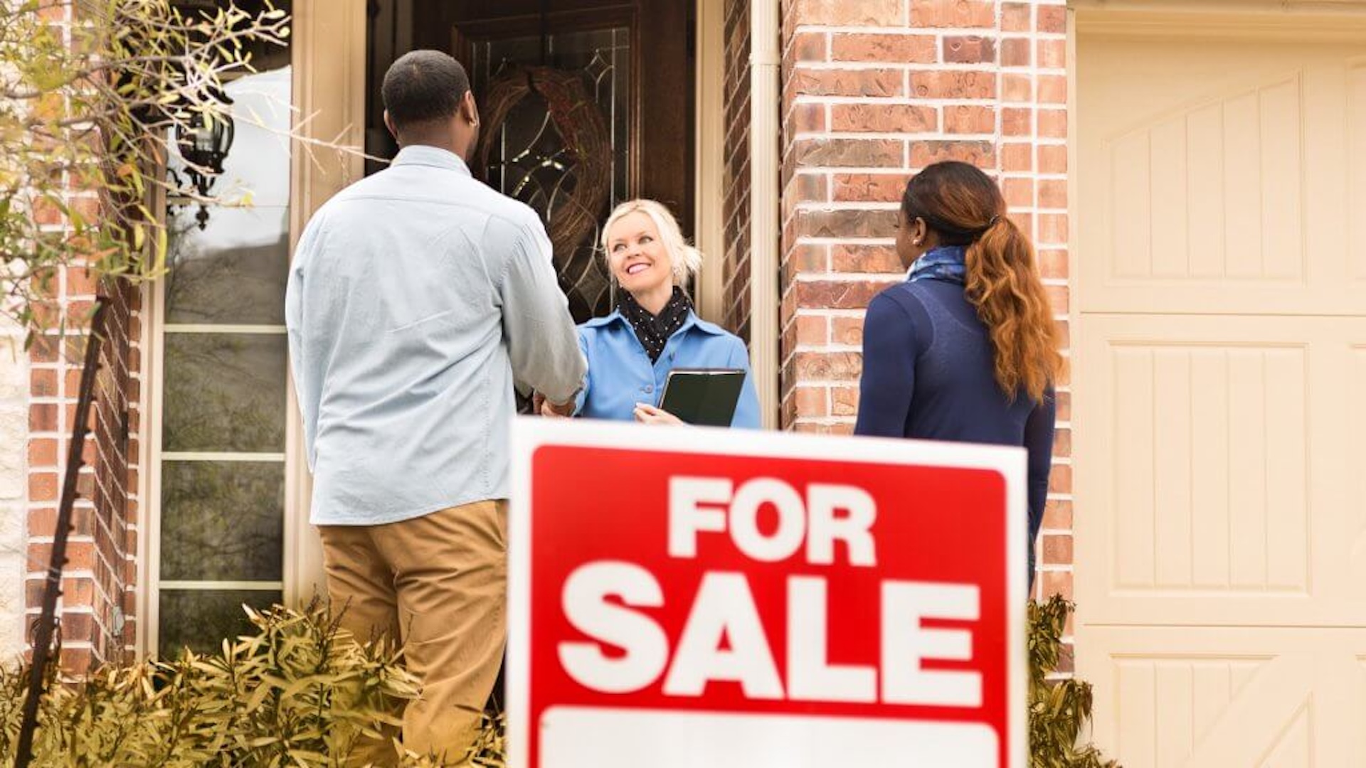 Discover a young couple shaking the hand of a smiling real estate agent at a house with a for sale sign.