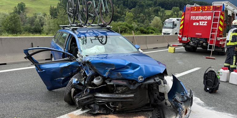 Schwer beschädigtes blaues Auto mit Fahrrädern auf dem Dach, Feuerwehr im Einsatz auf Autobahn.