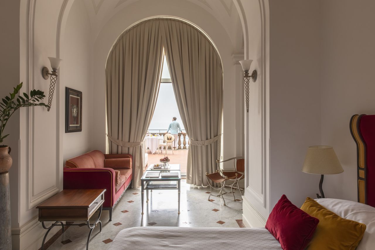 A luxurious guest room with a seating area a bed and a large window with drapes leading to a balcony where a person is standing