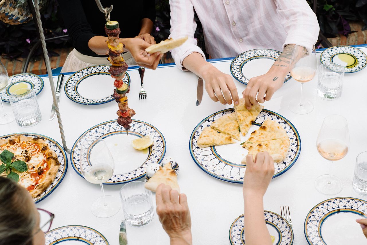A top view of a dining table with several people sharing food including plates of pizza and skewers