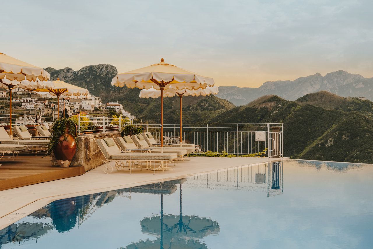 An infinity pool with lounge chairs and umbrellas on a patio overlooking hilly terrain and distant mountains