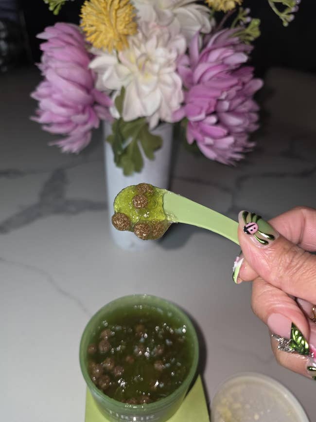 A person holds a spoon with brown round foods over a green smoothie; flowers are in the background