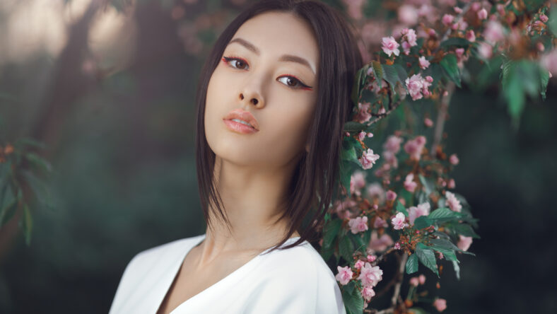 Asian woman fashion close-up portrait. Beautiful mixed race asian caucasian young girl perfect skin creartive art make-up wearing trendy white japanese kimono standing agaist flower background. Spring blossom of sakura tree in city park and elegant fashion lady.