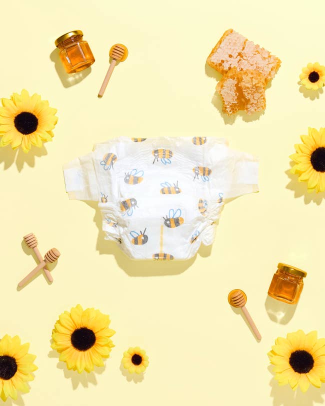 Baby diaper with bee print surrounded by honey jars, honey dippers, sunflower pieces, and honeycomb on a yellow background