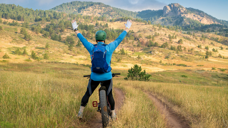 Back of a cyclist raising their arms in the air on a grassy path with mountains in the distance at Lory State Park