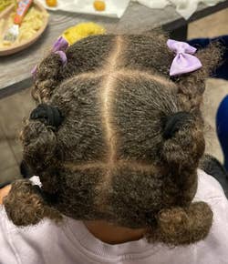 Child's hairstyle with four curly sections, two tied with purple bows and two with black ties, highlighting a creative and fun hairdo