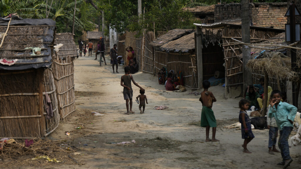 Children and adults on a dirt lane lined with thatched and bamboo dwellings in a rural South Asian village