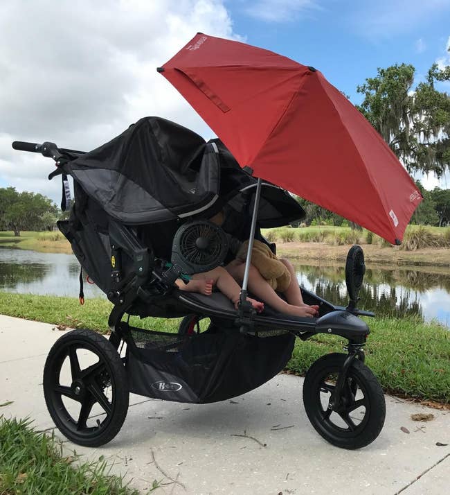 A double stroller with a large sunshade and a fan is positioned on a path by a pond, providing shade and comfort for two seated children