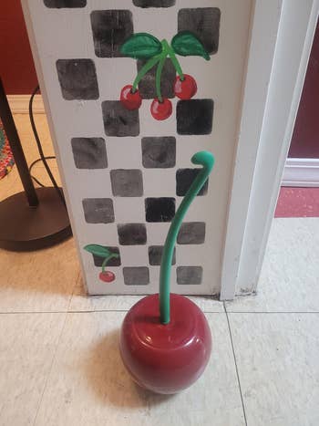 Whimsical cherry-themed toilet brush holder with green stem, red base. Cherry wall art in background. Featured in a shopping article