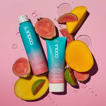 Sunscreen spray and lotion by COOLA, surrounded by mango and guava slices, arranged on a pink background