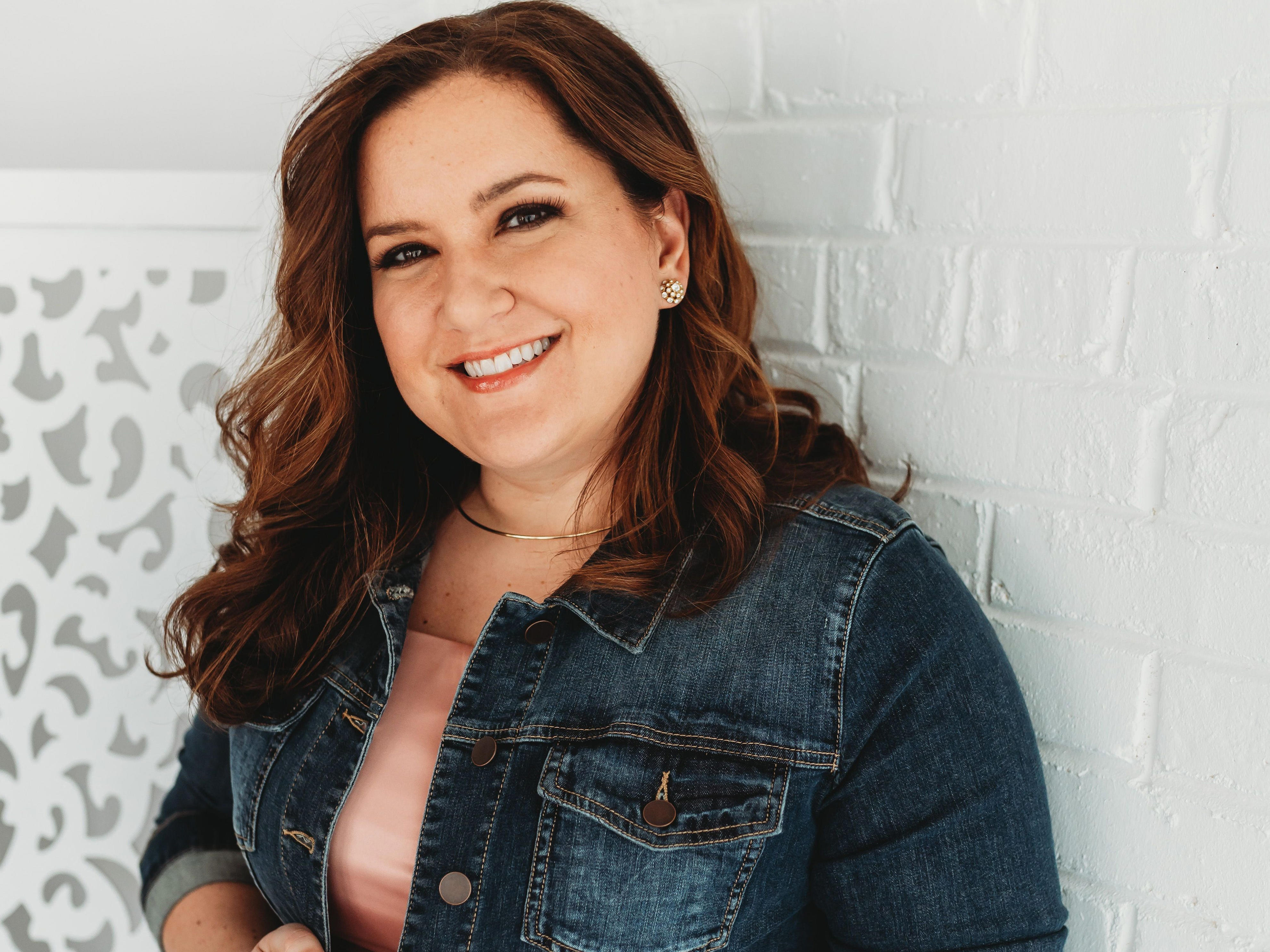 Smiling person in a denim jacket and pink dress poses against a white brick wall.