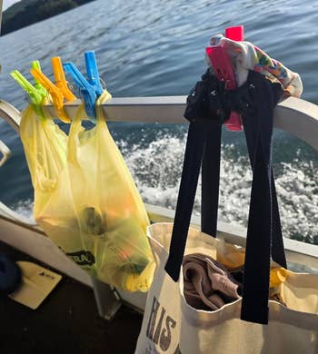 Tote bags and clothes hang from clips on a boat railing with water in the background