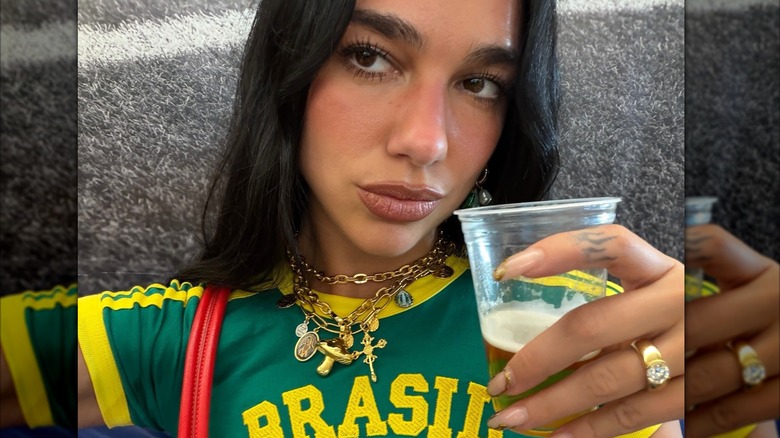 Selfie of Dua Lipa wearing a charm necklace and statement ring as she holds a beer glass