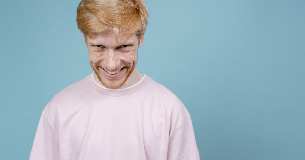 Young man in pink shirt smiles mischievously against blue background in studio setting.