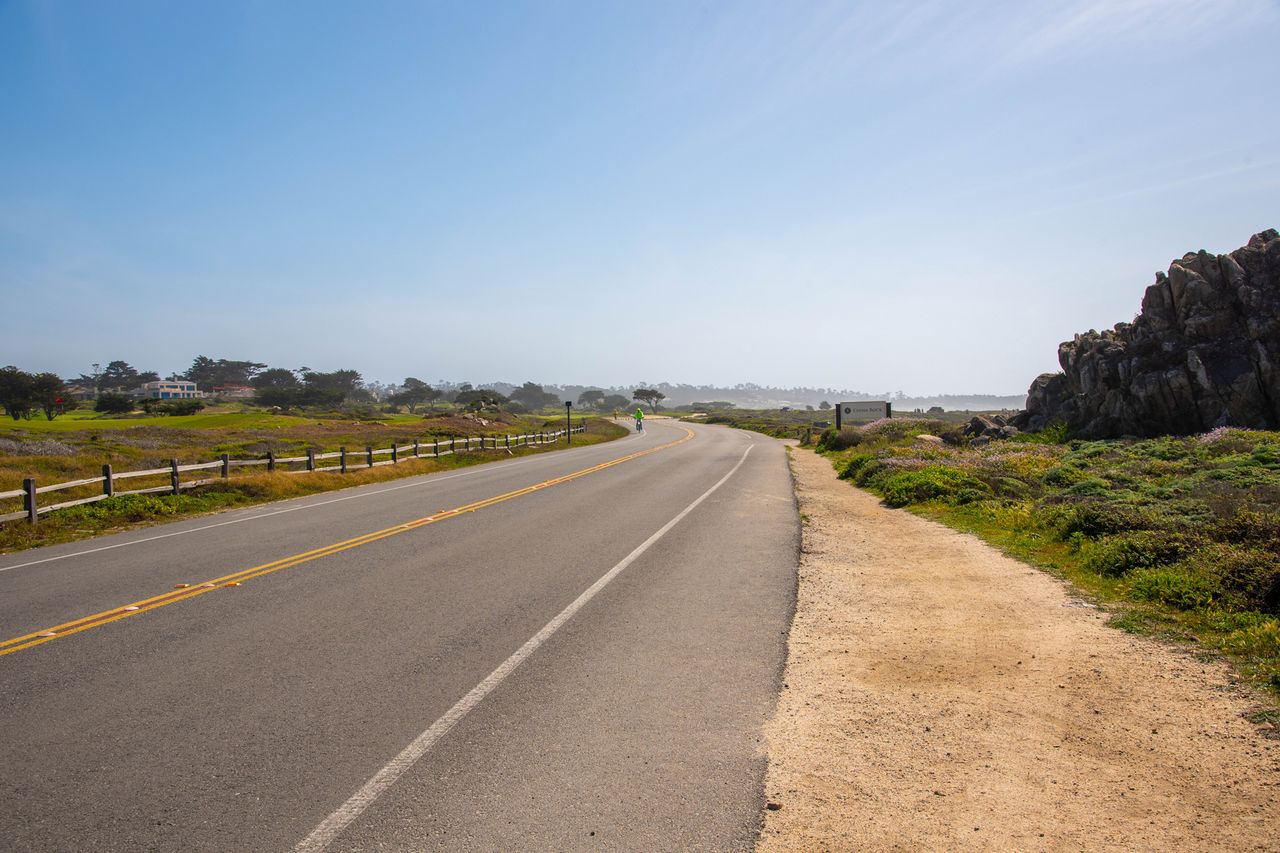 A paved road bordered by a wooden fence and greenery with flat terrain and hill formations in the background